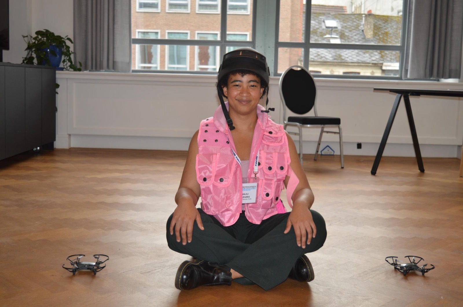 The artist seated between two drones in a pink tactical vest and helmet